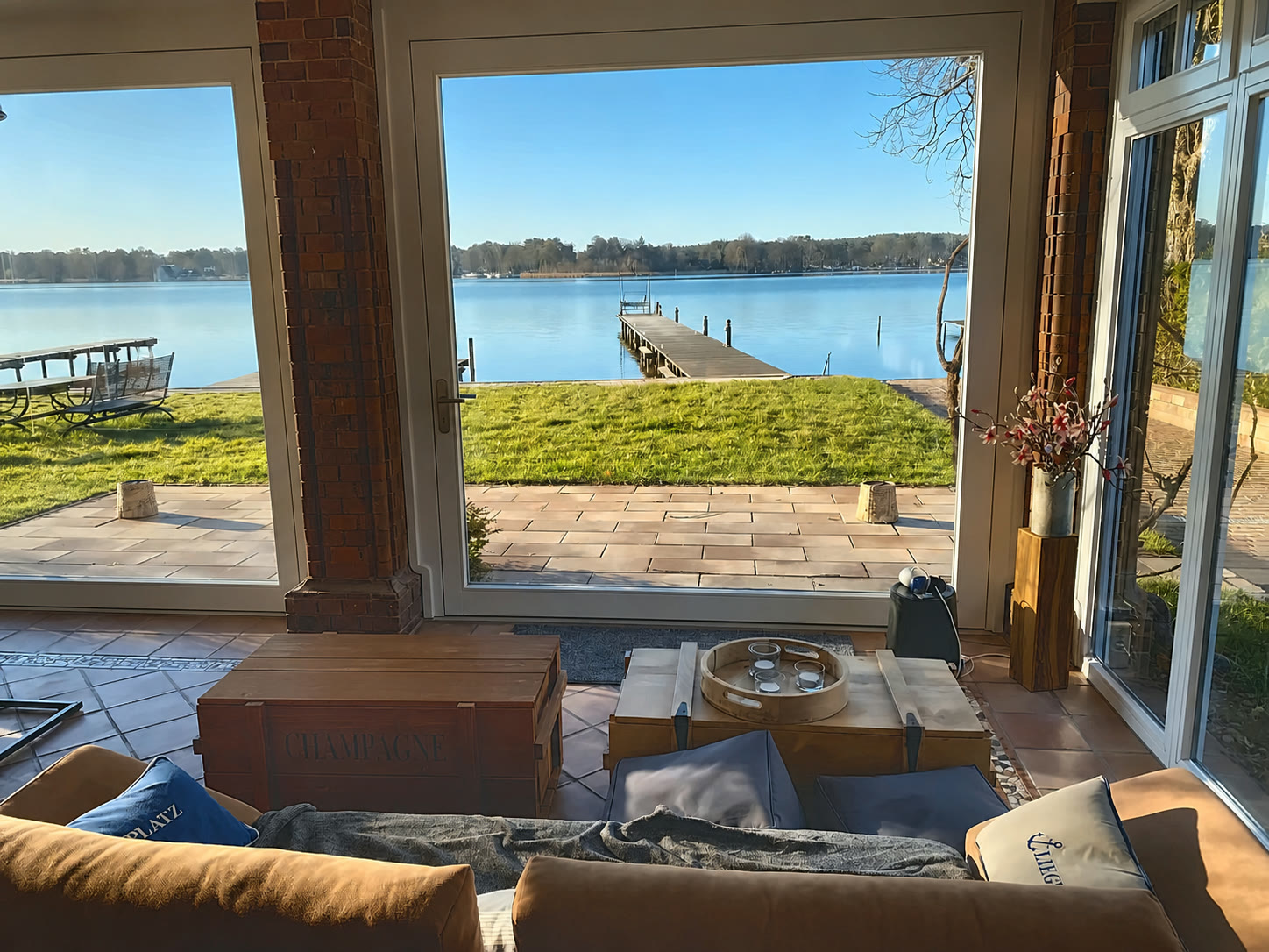 Wohnbereich mit bodentiefem Panoramafenster zum Krüpelsee — Blick über Terrasse und Uferwiese auf den Steg